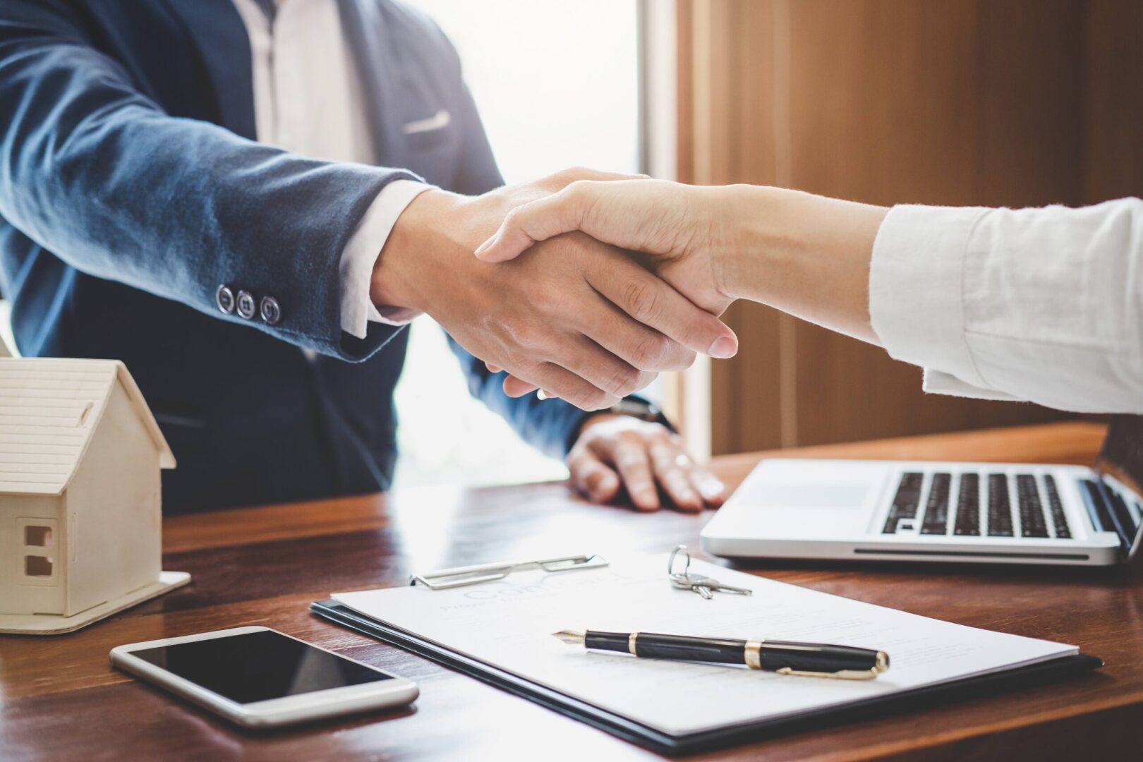 Two people shaking hands over a desk with papers and pens.